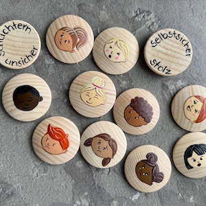 May include: A set of wooden discs featuring painted faces expressing different emotions. Some discs have German text, including "schüchtern unsicher" and "selbstsicher stolz." The faces display a variety of skin tones and hair colours, with expressions from joy to sadness.