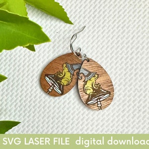 May include: A pair of wooden earrings with a laser-cut design of a frog wearing a hat and sitting on a mushroom. The earrings are brown with yellow, blue, and white accents.