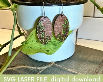 May include: A pair of oval-shaped wooden earrings with a green and brown leafy design. The earrings are hanging from a plant in a white pot.