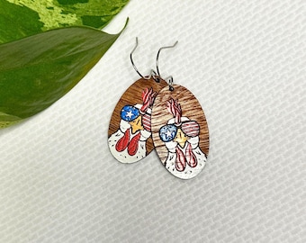 May include: A pair of wooden earrings featuring a hand-painted design of a rooster wearing sunglasses with an American flag pattern. The earrings are oval-shaped and have a brown wood base with a white and red painted design.