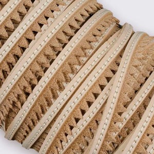 Puede incluir: Primer plano de un rollo de ribete decorativo de cuero. El ribete presenta un color marrón claro con un borde en zigzag y una tira de color crema en el centro, cosida con hilo blanco. El ribete se utiliza probablemente para proyectos de artesanía o costura.