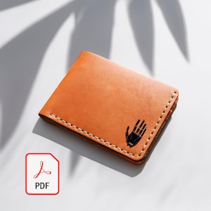Può includere: Un portafoglio in pelle color cuoio con una grafica a mano nera. Il portafoglio è rettangolare con cuciture bianche sui bordi. Il portafoglio è in pelle ed è uno stile a due ante.