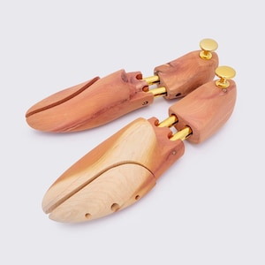 May include: A pair of wooden shoe trees crafted from light brown cedar. They feature a split-toe design and brass-coloured hardware. The heel has a round, gold-coloured knob. These shoe trees are designed to maintain shoe shape and absorb moisture, keeping shoes in good condition.