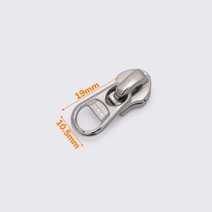 May include: A silver-colored zipper pull, measuring 19mm long and 10.5mm wide. The pull has a rounded design with a loop for easy gripping. The brand name "YKK" is visible on the pull.