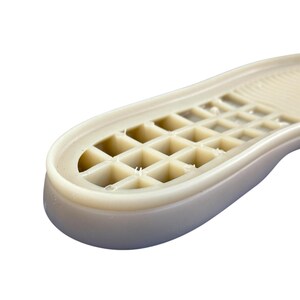 May include: Close-up of a beige shoe sole with a grid-like pattern. The sole has a rounded toe and a textured surface, designed for grip and support. The material appears to be a durable, synthetic rubber.