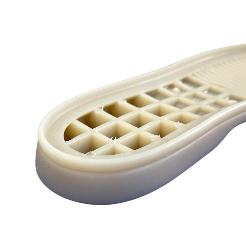May include: Close-up of a beige shoe sole with a grid-like pattern. The sole has a rounded toe and a textured surface, designed for grip and support. The material appears to be a durable, synthetic rubber.