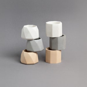 May include: A set of six geometric concrete planters in various shades of grey, white, and beige. The planters are stacked in a pyramid shape.