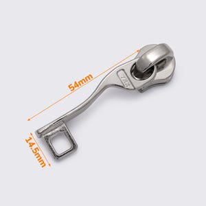 Op de afbeelding: Zilveren rits-trekker met een 13,7 cm lange handgreep en een 3,7 cm brede basis. De trekker heeft een kleine vierkante lus aan het einde van de handgreep en een ronde, draaibare knop aan de bovenkant. De knop heeft de tekst "ZKK" erop gedrukt.