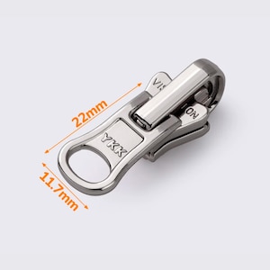 Puede incluir: Tirador de cremallera YKK plateado con asa de bucle, que mide 22 mm de largo y 1,7 mm de ancho. El tirador tiene las palabras "YKK" y "VISLON" impresas.