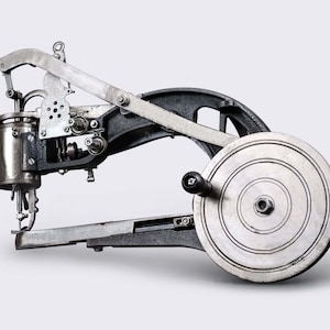 May include: A vintage industrial sewing machine with a large metal wheel and a lever arm. The machine is made of metal and has a silver finish.
