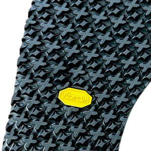 Op de afbeelding: Close-up van een zwarte rubberen zool met een textuurpatroon van kruisvormen. Een geel "Vibram"-logo is zichtbaar. De zool is ontworpen voor schoeisel en biedt grip en duurzaamheid.