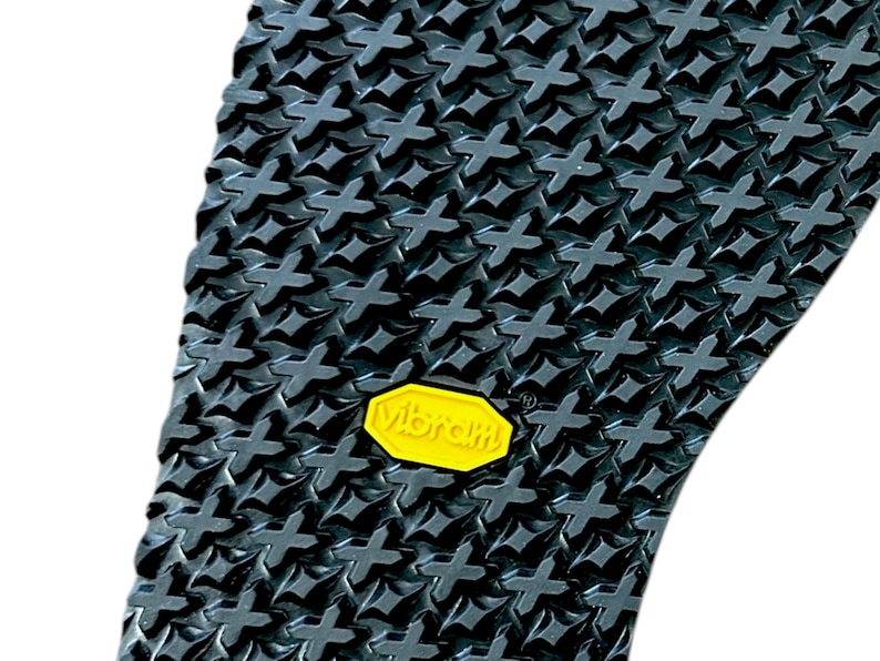 Op de afbeelding: Close-up van een zwarte rubberen zool met een textuurpatroon van kruisvormen. Een geel "Vibram"-logo is zichtbaar. De zool is ontworpen voor schoeisel en biedt grip en duurzaamheid.