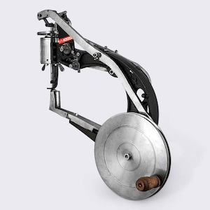 May include: A vintage industrial sewing machine with a large metal wheel and a black metal arm. The machine is designed for sewing leather and other heavy materials.