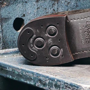 K&ouml;nnte beinhalten: Nahaufnahme der Sohle eines braunen Lederschuhs mit einer schwarzen Gummisohle. Die Sohle hat ein Muster aus kleinen Kreisen und einem gr&ouml;&szlig;eren Kreis mit dem Text "Crockett & Jones" und "Made in England".