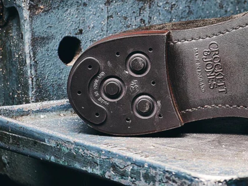 K&ouml;nnte beinhalten: Nahaufnahme der Sohle eines braunen Lederschuhs mit einer schwarzen Gummisohle. Die Sohle hat ein Muster aus kleinen Kreisen und einem gr&ouml;&szlig;eren Kreis mit dem Text "Crockett & Jones" und "Made in England".