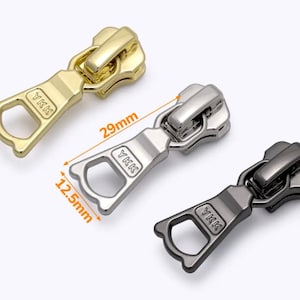 Puede incluir: Tres tiradores de cremallera en metal dorado, plateado y negro. Cada tirador mide 29 mm de largo y 12,5 mm de ancho. Los tiradores tienen el logotipo de YKK.