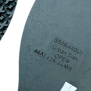 Op de afbeelding: Close-up van een grijze schoenzool met de tekst "S509(440C) Urban Trek OPEN 44A(42A-44A)R". De zool heeft een getextureerd oppervlak en een patroon op de rand.