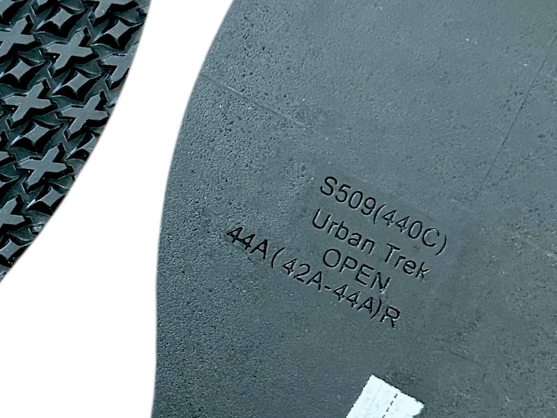 Op de afbeelding: Close-up van een grijze schoenzool met de tekst "S509(440C) Urban Trek OPEN 44A(42A-44A)R". De zool heeft een getextureerd oppervlak en een patroon op de rand.