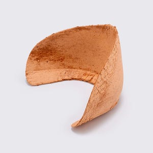 Peut inclure: Un bracelet manchette incurvé et ouvert, en matériau brun rougeâtre texturé. Le bracelet a une forme arrondie avec un intérieur lisse et un extérieur texturé. Il est présenté sur un fond blanc.