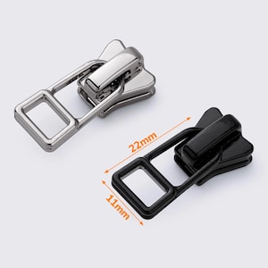 Op de afbeelding: Twee ritsen, een zilveren en een zwarte, met een rechthoekige lus. De zilveren rits is 22 mm lang en 11 mm breed. De zwarte rits is even groot.