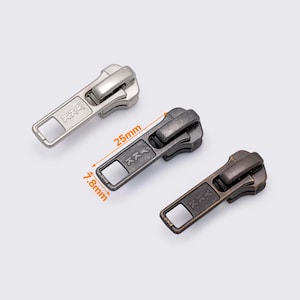 May include: Three metal zipper pulls in silver, gunmetal, and bronze. The gunmetal zipper pull is 2.5cm long and 0.78cm wide. Each pull has a rectangular handle with a square opening and a decorative design.
