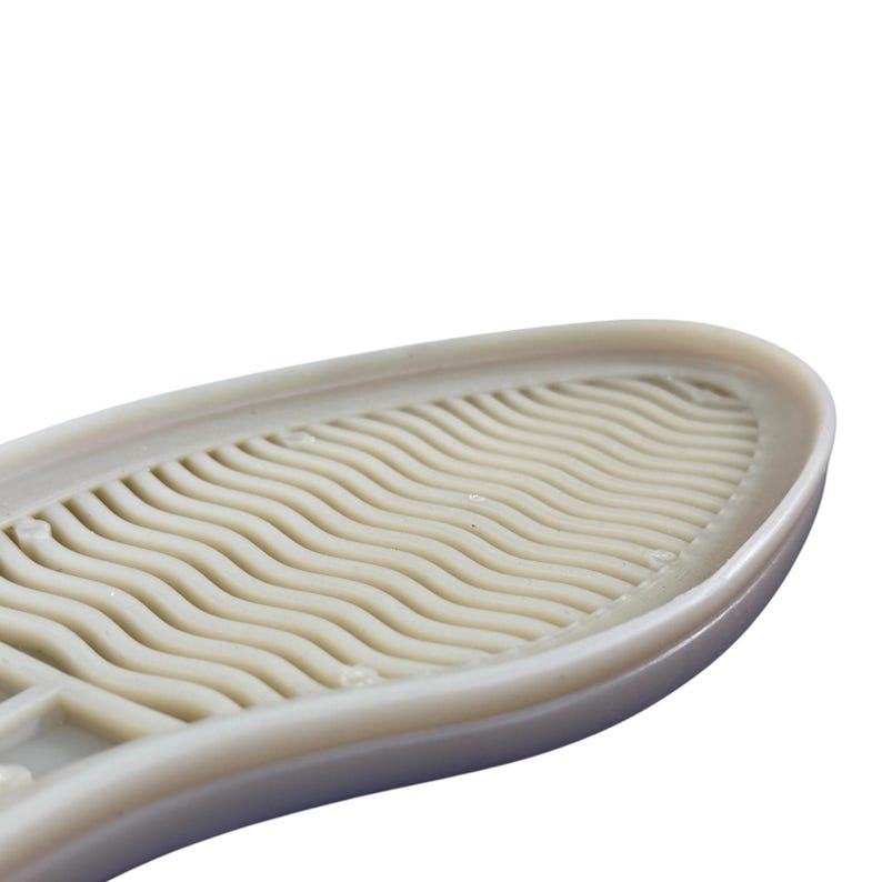 May include: A close-up of a beige shoe sole, featuring a wavy, textured pattern. The oval-shaped sole is made of a flexible material. The design suggests a focus on grip and comfort for footwear.