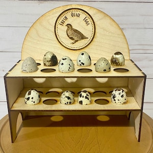 Op de afbeelding: Een houten eierrek met de tekst "Fresh Quail Eggs" en een afbeelding van een kwartel. De rek heeft twee lagen met 12 vakjes voor eieren. De rek is gemaakt van lichtbruin hout en heeft een natuurlijke afwerking.