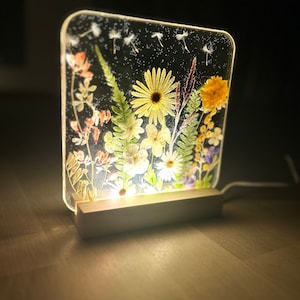 May include: A square, clear resin night light with a white base. The resin contains pressed flowers and greenery, including yellow daisies, purple wildflowers, and green ferns. The light is illuminated, casting a warm glow.