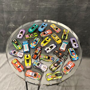 May include: A clear glass table with a black metal base, decorated with a variety of colorful toy cars. The cars are arranged in a circular pattern on the table's surface.