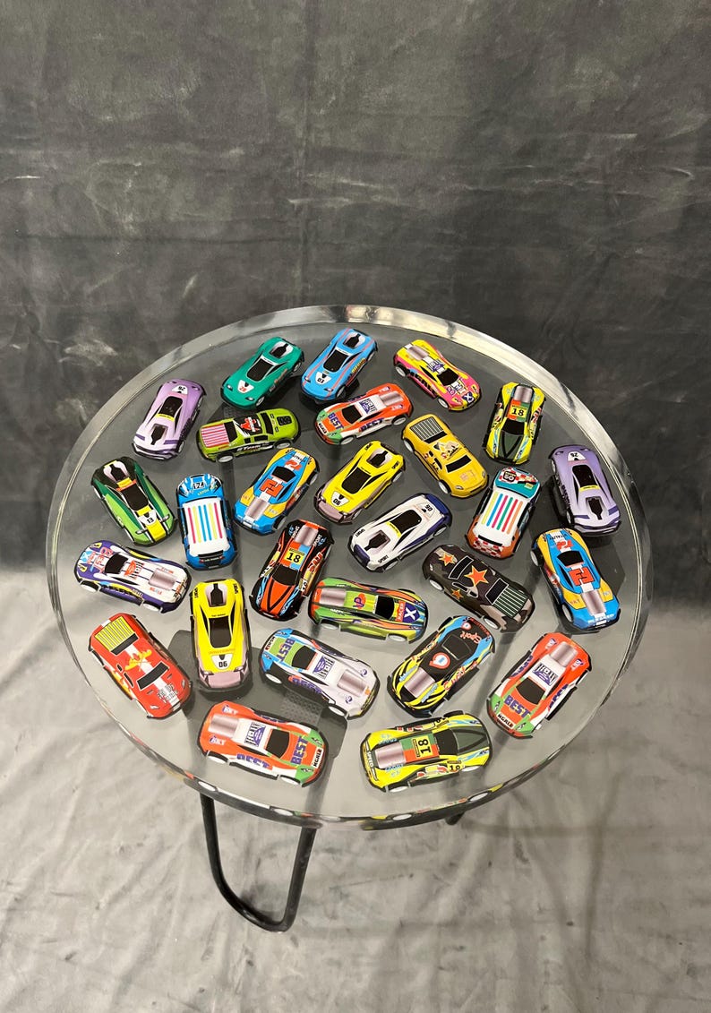 May include: A clear glass table with a black metal base, decorated with a variety of colorful toy cars. The cars are arranged in a circular pattern on the table's surface.