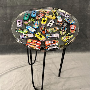 May include: A round table with a clear resin top filled with colorful toy race cars. The table has a black metal base with three hairpin legs.