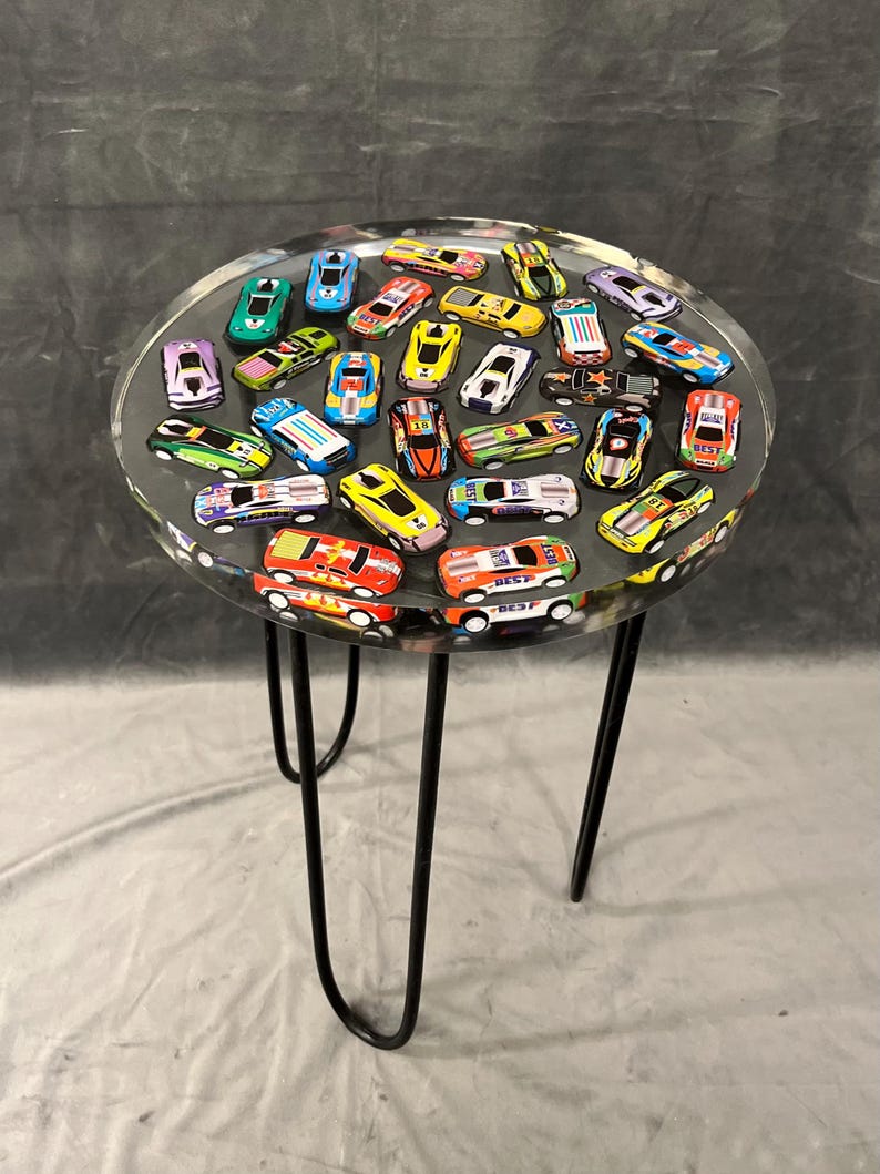 May include: A round table with a clear resin top filled with colorful toy race cars. The table has a black metal base with three hairpin legs.
