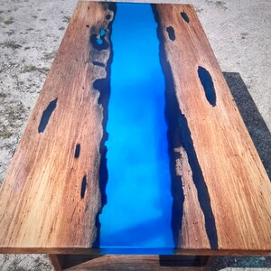 Op de afbeelding: Een rechthoekige houten tafel met een levendige blauwe epoxyharsrivier in het midden. Het hout heeft een natuurlijke nerf en donkere accenten. De tafel is een uniek meubelstuk.