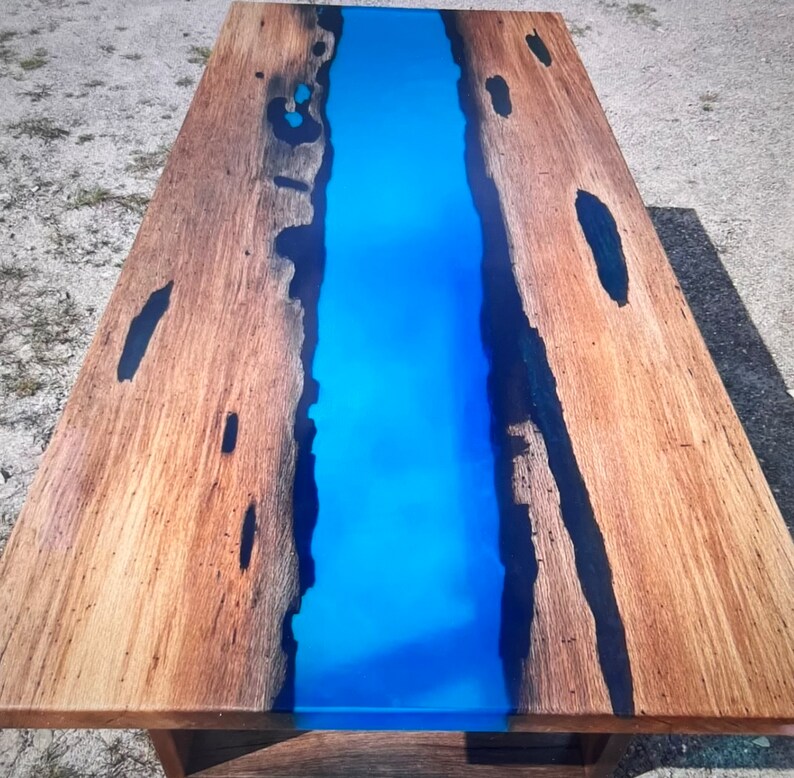Op de afbeelding: Een rechthoekige houten tafel met een levendige blauwe epoxyharsrivier in het midden. Het hout heeft een natuurlijke nerf en donkere accenten. De tafel is een uniek meubelstuk.