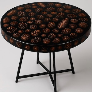 Peut inclure: Une table ronde noire avec un piètement en métal noir. Le plateau de la table a un motif de pommes de pin brunes sur fond noir.