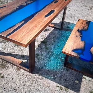Op de afbeelding: Een houten tafel en een bijpassende bijzettafel met een blauw epoxyhars rivierontwerp. De tafels hebben een natuurlijke houtnerf afwerking en donkerbruine poten, wat een moderne, artistieke uitstraling cre&euml;ert. Unieke meubels.