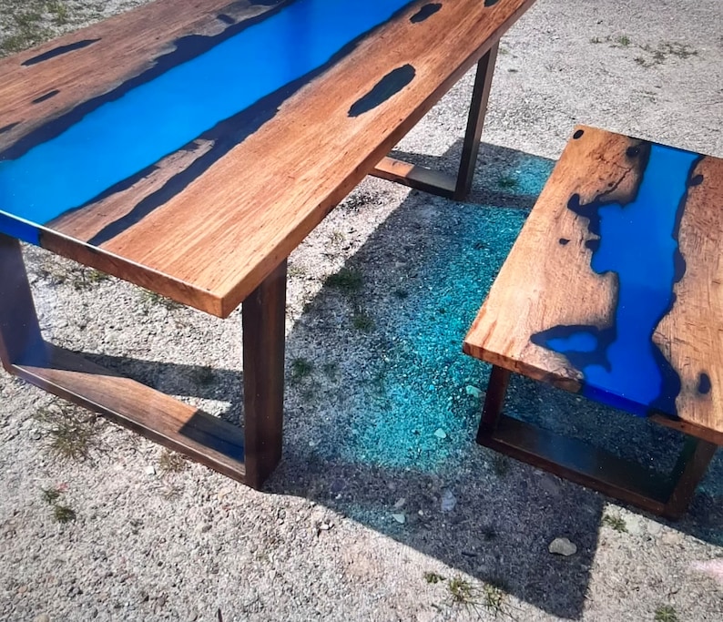 Op de afbeelding: Een houten tafel en een bijpassende bijzettafel met een blauw epoxyhars rivierontwerp. De tafels hebben een natuurlijke houtnerf afwerking en donkerbruine poten, wat een moderne, artistieke uitstraling cre&euml;ert. Unieke meubels.