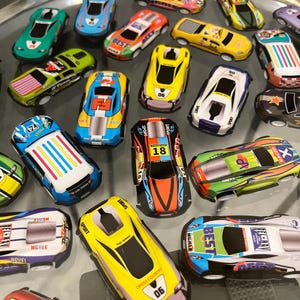 May include: A collection of colorful toy race cars with various designs and numbers. The cars are made of plastic and have a variety of colors, including red, blue, green, yellow, and black. The cars are all different sizes and shapes, and some have different designs on them. The cars are all on a clear surface.