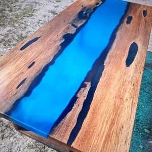 Op de afbeelding: Een rechthoekige houten salontafel met een levendige blauwe epoxyharsrivier in het midden. Het hout heeft een natuurlijke nerf en donkere accenten. De tafel is ongeveer 152 cm lang en 76 cm breed.