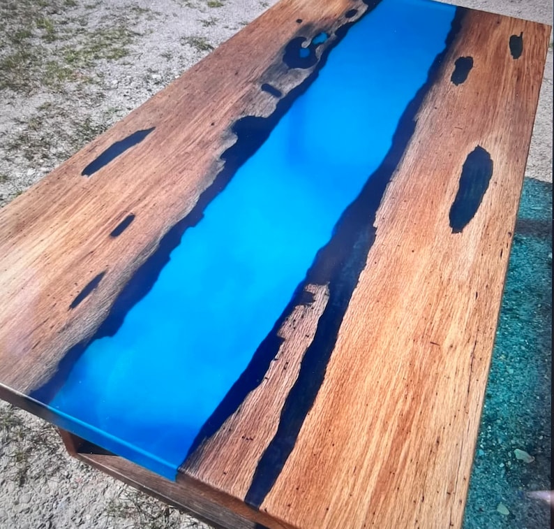 Op de afbeelding: Een rechthoekige houten salontafel met een levendige blauwe epoxyharsrivier in het midden. Het hout heeft een natuurlijke nerf en donkere accenten. De tafel is ongeveer 152 cm lang en 76 cm breed.