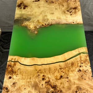 May include: A wooden table top with a green epoxy river running through it. The wood has a natural burl pattern and is a light brown color.