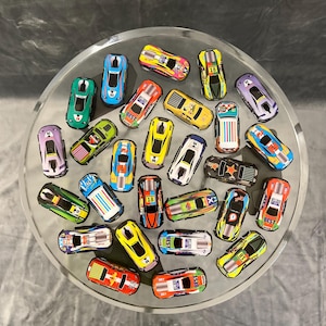 May include: A collection of 25 toy race cars in various colors and designs. The cars are arranged in a circle on a silver surface. Some of the cars have numbers on them, such as "18" and "50".