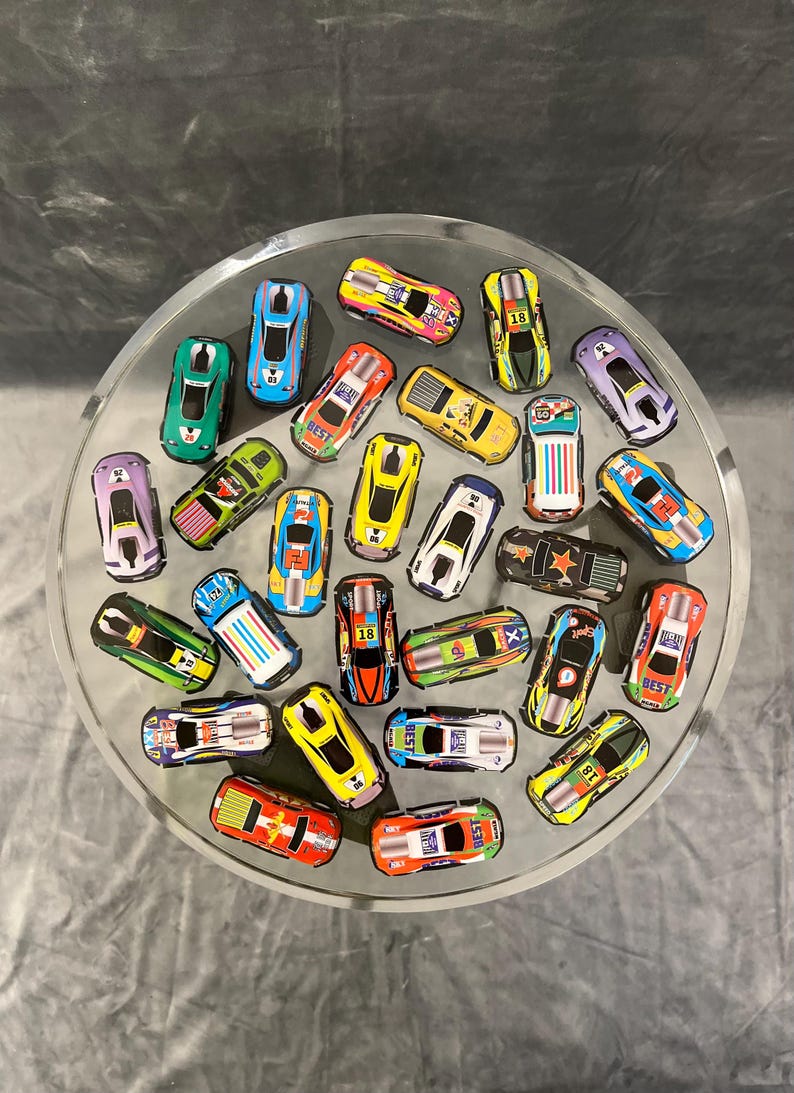 May include: A collection of 25 toy race cars in various colors and designs. The cars are arranged in a circle on a silver surface. Some of the cars have numbers on them, such as "18" and "50".
