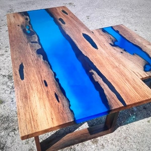 Op de afbeelding: L-vormige houten tafel met een levendige blauwe epoxyharsrivier in het midden. De tafel heeft een natuurlijke houtnerf afwerking met donkere accenten en een donkerbruine basis. De tafel is ongeveer 152 cm lang en 76 cm breed.
