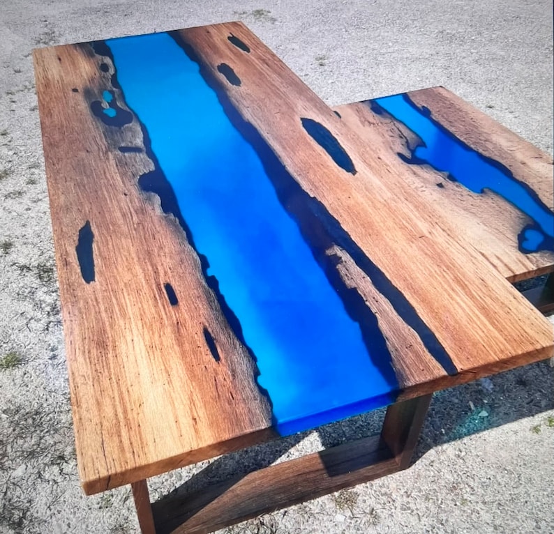 Op de afbeelding: L-vormige houten tafel met een levendige blauwe epoxyharsrivier in het midden. De tafel heeft een natuurlijke houtnerf afwerking met donkere accenten en een donkerbruine basis. De tafel is ongeveer 152 cm lang en 76 cm breed.