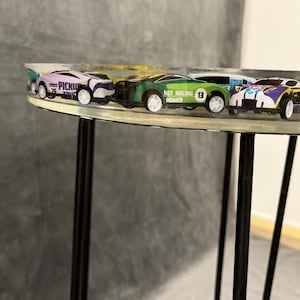 May include: A clear round table with black metal legs.  Five toy cars are embedded in the table top.  The cars are purple, green, white, and blue.  One car has the text "Hot Racing Power 13" on it.