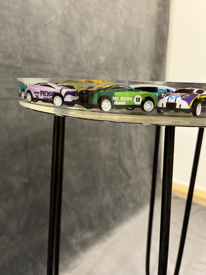 May include: A clear round table with black metal legs.  Five toy cars are embedded in the table top.  The cars are purple, green, white, and blue.  One car has the text "Hot Racing Power 13" on it.