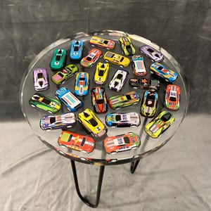 May include: A clear glass table top with a black metal base. The table top is filled with colorful toy race cars. The cars are all different colors and designs, and some have numbers on them.