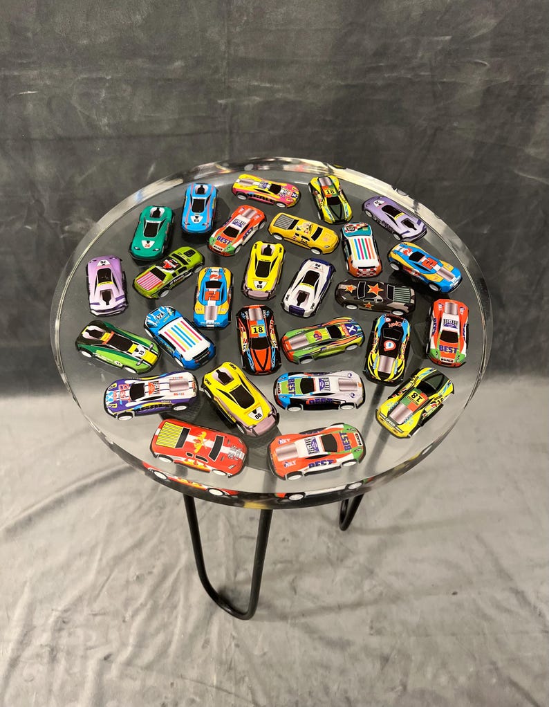 May include: A clear glass table top with a black metal base. The table top is filled with colorful toy race cars. The cars are all different colors and designs, and some have numbers on them.