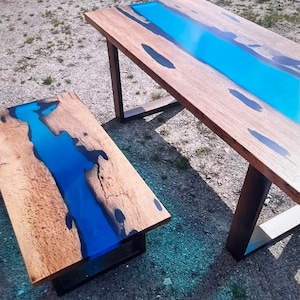 Op de afbeelding: Twee houten tafels met een blauw hars rivierontwerp. De grotere tafel heeft een rechthoekige vorm met donkere metalen poten. De kleinere tafel is een salontafel met een vergelijkbaar ontwerp en metalen voet. Het hout heeft een natuurlijke nerf.